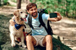 A male hiker with a backpack and a labrador dog makes a selfie on the phone while sitting on a stone in the forest. Camping, travel, hiking.