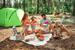 Happy family with a child and a dog at a picnic sit on a blanket near the tent and eat fried food and watermelon during the weekend in the forest. Camping, recreation, hiking.