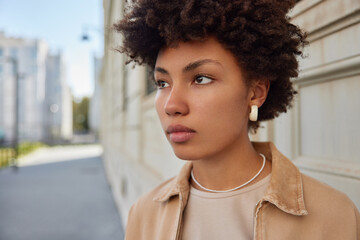Wall Mural - Close up shot of good looking thoughtful woman with Afro hair focused into distance with serious expression spends free time outside thinks about future plans. People leisure lifestyle concept