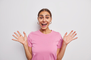 Wall Mural - Happy cheerful woman raises palms feels glad smiles cheerfully reacts on amazing news dressed in casual pink t shirt isolated over white background. People human reactions and feelings concept