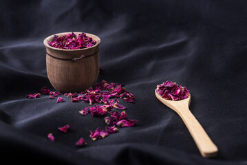 Wall Mural - closeup shot of dry rose petals in a wooden pot with a wooden spoon on a black fabric