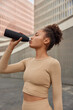Slim active woman feels thirsty drinks fresh water from bottle dressed in sportsclothes takes break during workout poses in downtown. Female runner hydrates after training leads healthy lifestye
