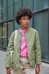 Wall Mural - Serious beautiful curly haired woman wears stylish jacket trouses poses outside with pensive expression looks away discovers new places of interest during excursion time. Recreation concept.