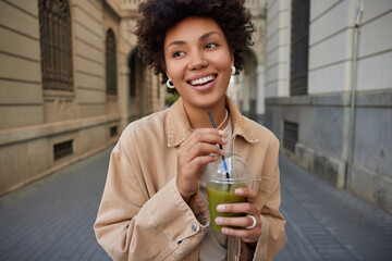Wall Mural - Glad carefree Afro American woman drinks smoothie from straw looks gladfully somewhere enjoys recreation time feels happy poses in ancient city enjoys good day. Positive female with refreshing drink