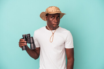 Wall Mural - Young African American man holding binoculars isolated on blue background confused, feels doubtful and unsure.