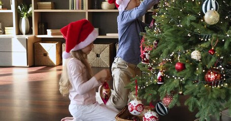 Wall Mural - Preparing for New Year. Happy friendly children elder brother younger sister in Santa caps decorate Christmas tree with big glitter balls toys. Preteen siblings enjoy dressing fir tree at living room