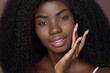 Young black African woman model with curly afro hair touching healthy flawless radiant smooth glowing face skin care isolated on brown background. Ethnic natural beauty skincare concept. Portrait