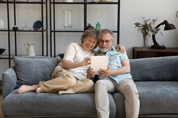 Happy bonding mature middle aged family couple using digital computer tablet, enjoying playing online games, web surfing information, choosing goods shopping in internet store, tech addiction concept.