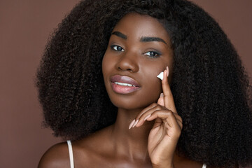 Beautiful happy African American young woman with afro hair and perfect smooth skin complexion applying putting creme on face doing daily routine. Skincare, cosmetology and spa concept. Close up.