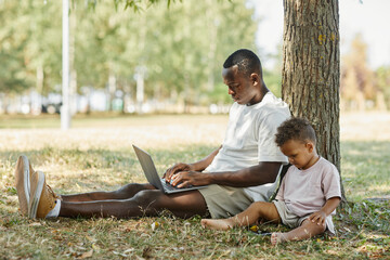 Wall Mural - Side view portrait of young African-American man using laptop outdoors in park with cute baby son, copy space