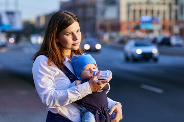 Woman is feeding child in baby sling from bottle near traffic.