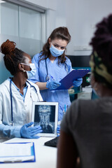 Wall Mural - African american therapist showing medical radiography to sick patient discussing healthcare treatment with asisstant during examination in hospital office. People with face mask against coronavirus