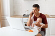 © sementsova321 - Indoor shot of man wearing burgundy casual t shirt with towel on his shoulder, looking after baby and working online from home, talking phone and giving water to his kid.