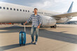Smiling Caucasian traveler posing with travel bag with while plane on the background. Trip concept