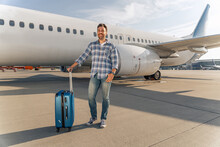 Smiling Caucasian Traveler Posing With Travel Bag With While Plane On The Background. Trip Concept