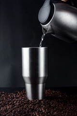 Wall Mural - Hand holding electric Kettle and pouring water into stainless steel tumbler of hot black coffee place on pile of coffee seed on black backgroun.