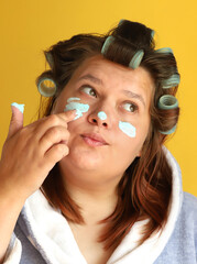 A plus size model gir in curlers and a dressing gown in the process of applying a cosmetic face mask