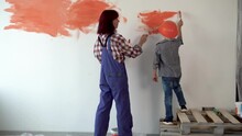 A Woman Standing Next To A Child Applies Orange Paint To The Wall In Her Home With A Paint Brush With Her Back To The Camera.
