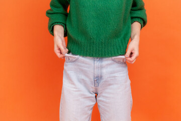 Unknown faceless woman wearing green casual style sweater showing empty pockets, has mo money, worried about debts and bankruptcy. Indoor studio shot isolated on orange background.