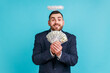 © khosrork - Happy businessman wearing official style suit with halo over head holding fan of dollar bills and smiling happily, showing money to camera. Indoor studio shot isolated on blue background.
