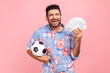 © khosrork - Extremely excited happy man with beard in blue casual shirt holding soccer ball and dollar bills, screaming, betting and winning, keeps eyes closed. Indoor studio shot isolated on pink background.