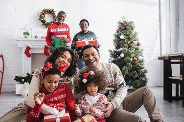 happy african american family with gift boxes looking at camera near blurred grandparents and christmas tree