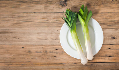 Two fresh raw leeks lie on a white plate on an old wooden table. View from above. Copy space