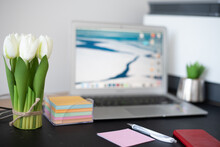 Work Business Place Of Woman. White Artificial Flowers Of Tulips On Background Open Blurred Laptop. Women's Organized Black Table. Pen And Notes. Don't Forget Anything. Order In The Workplace.