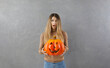 © Studio Romantic - Portrait of funny surprised and little scared teen girl standing with pumpkin in her hands. Girl in casual clothes posing with Halloween pumpkin standing on gray background. Halloween concept. Banner.