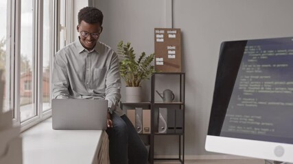 Medium stab shot of young male African-American programmer sitting on windowsill with laptop writing program codes appearing on monitor screen in modern office