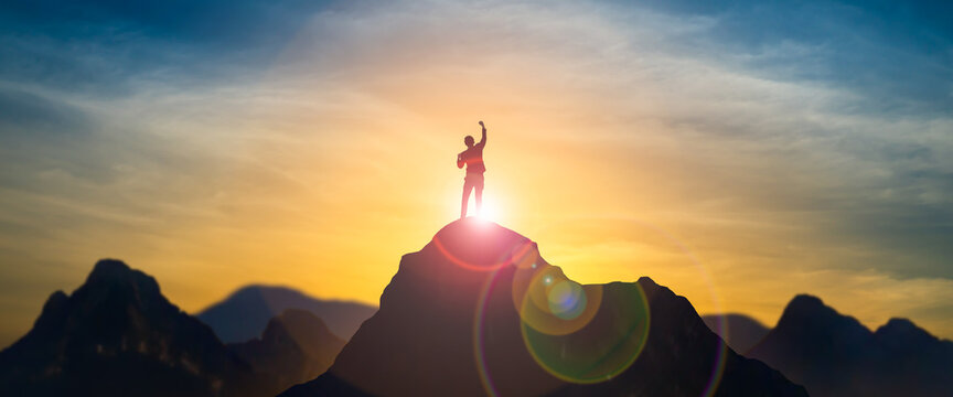 silhouette of businessman celebrating raising arms on the top of mountain with over blue sky and sun