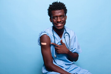 African american nurse pointing to vaccine shot bandage while looking at camera and smiling. Black medical assistant showing adhesive plaster after getting vaccinated against coronavirus