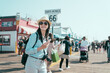 end of route 66 on santa monica pier california. happy smiling young asian chinese woman backpacker holding smartphone and camera joyful looking aside under sunshine. pretty tourist enjoy sun flare