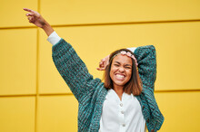 Happy Young Woman With Hand Raised Standing In Front Of Yellow Wall