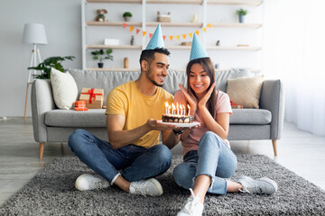 Poster - Happy millennial couple wearing party hats, celebrating birthday with festive cake at home, full length