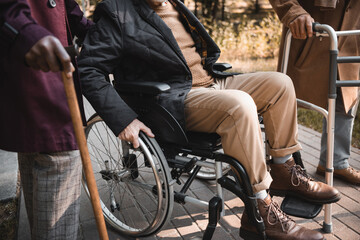 Wall Mural - Cropped view of man in wheelchair near interracial friends with walking frame and cane in park