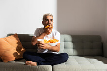 Hispanic Man Eating Candy On A Couch While Whatching Television. Unhealthy Diet And Bad Habits Concept.