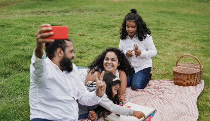 Happy indian family enjoy day outdoor at park with picnic and toys while taking a selfie using mobile phone - Family, parents and children love