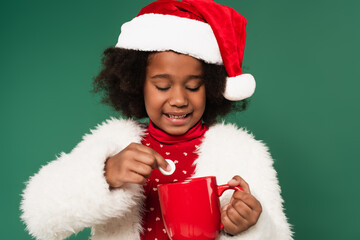 Canvas Print - Happy african american child in santa hat and fluffy jacket holding marshmallow near cup isolated on green