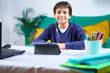 Smart and cheerful child looking at camera in online classes with tablet and laptop sitting on the couch at home