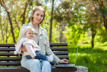 Stylish Business Mom Uses A Laptop With A Baby In Her Arms While Sitting On A Bench In The Park. Distance Work, Motherhood, Work On Maternity Leave