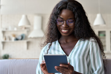 Happy black mixed raced African black young woman in glasses with perfect smile sitting on sofa holding using tablet device reading e book, surfing social media dating apps in apartment.