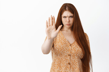 Stay back. Serious young redhead woman extend one hand to stop you, showing prohibit gesture, standing over white background