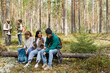 © Seventyfour - Full length portrait of diverse group of friends taking break while enjoying hike in forest, focus on three people sitting on log, copy space