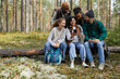 © Seventyfour - Full length portrait of diverse group of friends sitting on log in forest and using smartphone while enjoying hiking in Autumn, copy space
