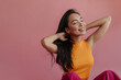 Studio shot of long-haired asian model smiling with her eyes closed and ruffling hair. Young woman is sitting on floor on pink background in orange and pink clothes.