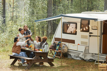 Wide angle view at diverse group of young people clinking beer bottles while enjoying picnic outdoors at campsite with trailer van