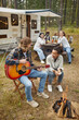 © Seventyfour - Vertical full length portrait of young couple playing guitar while camping with friends in forest
