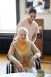 Vertical portrait of young woman assisting smiling senior woman in wheelchair at nursing home