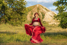 A Young Beautiful Red-haired Girl In A Red Ball Gown Is Dancing A Fiery Dance On A Green Meadow Against The Backdrop Of A Relict Mountain.